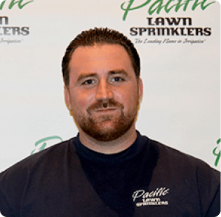 A man with short dark hair and a beard is standing in front of a backdrop that says "Pacific Lawn Sprinklers". He is wearing a navy blue shirt with the same company's logo on it.