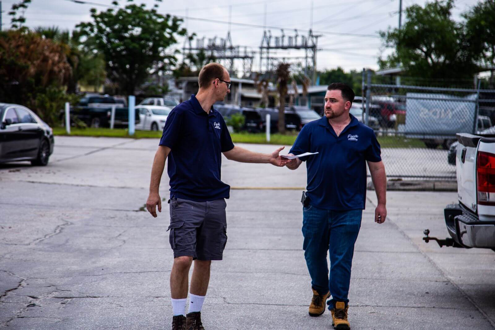 Two men standing outdoors, one handing a paper to the other. Both are wearing dark blue shirts and work boots. They are in a parking area with cars, a chain-link fence, and trees in the background.