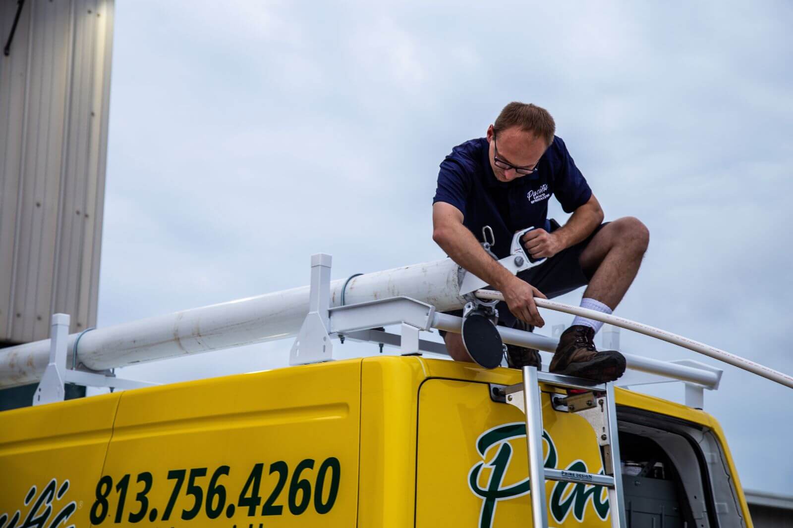 A man in a dark shirt is installing or adjusting a long white pipe on the roof of a yellow vehicle with a company name and contact number printed on the side.