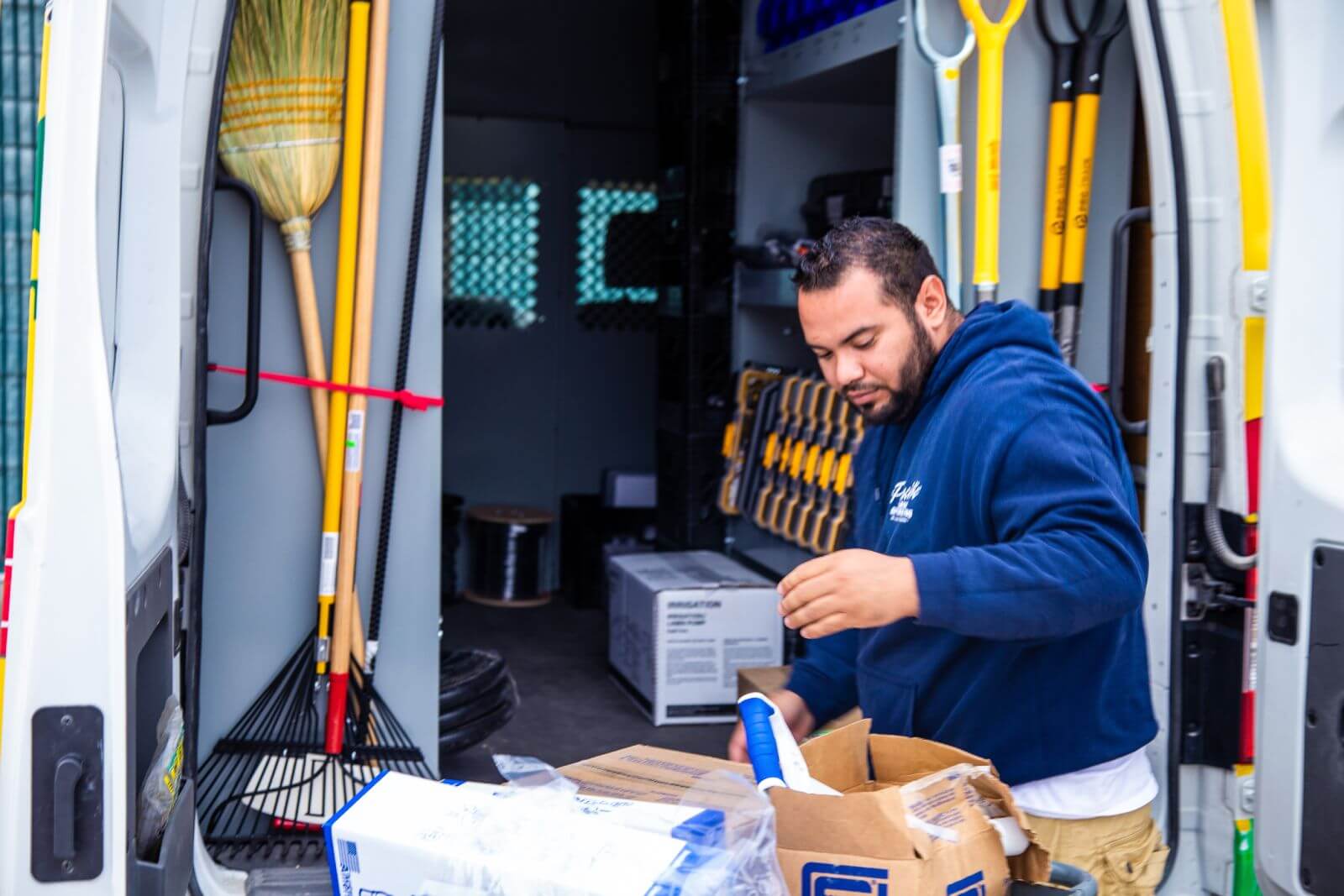 A man in a blue hoodie stands inside a van unpacking boxes, surrounded by various tools including brooms and rakes.