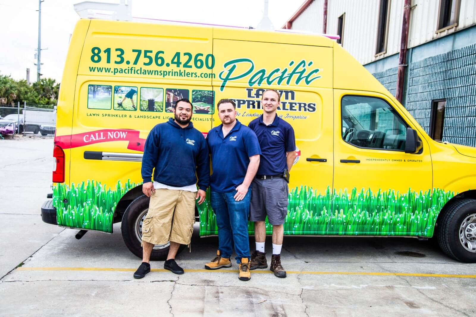 Three men standing in front of a yellow and green "Pacific Lawn Sprinklers" van parked by a building. The van features contact details and images related to lawn sprinkling services.