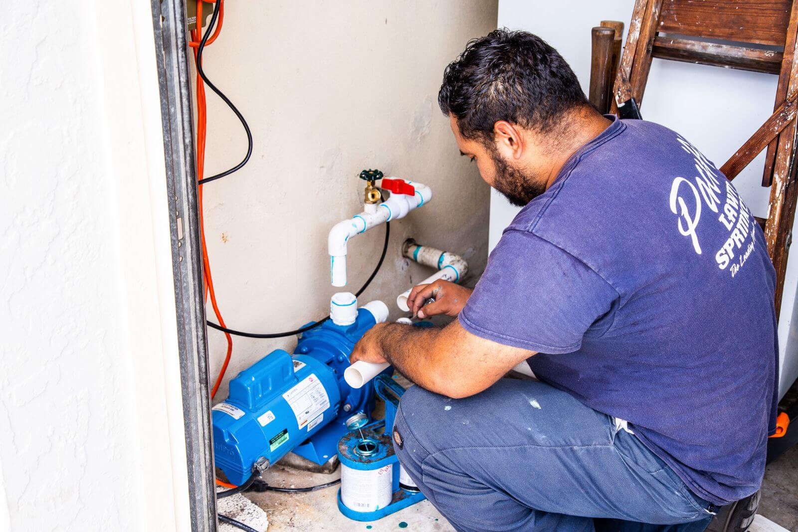 A man is kneeling and fixing pipes connected to a blue water pump in a small utility space, surrounded by tools and equipment.