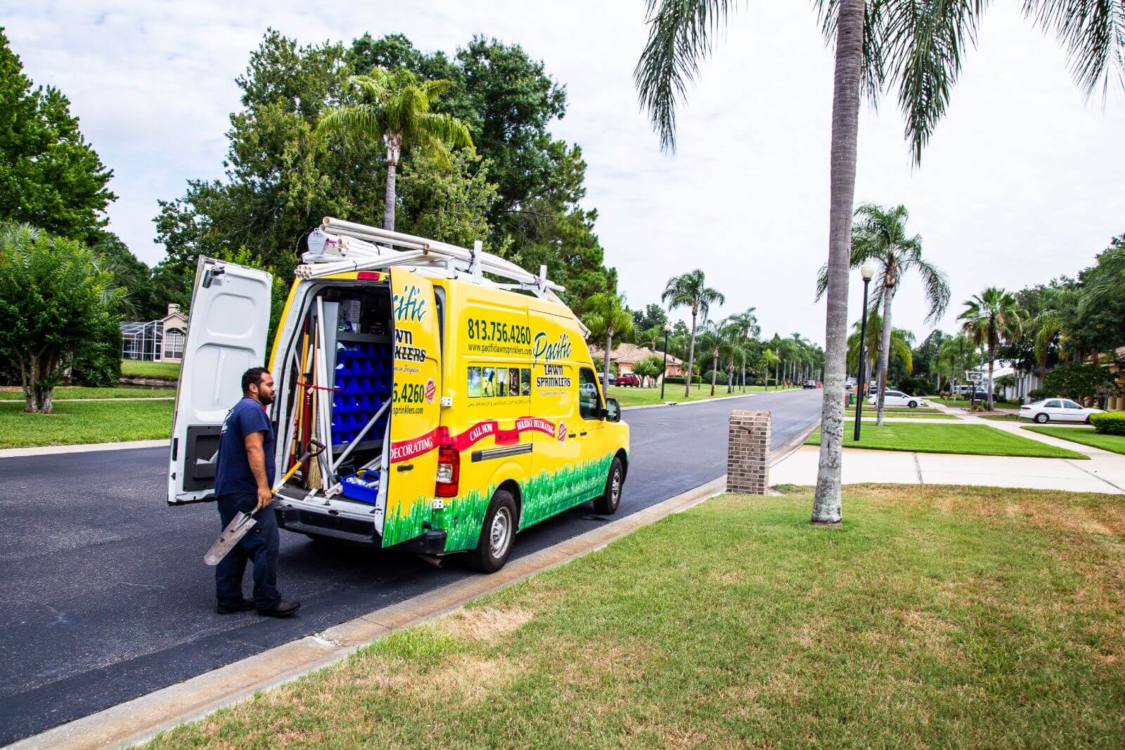 A man stands beside a yellow and green van with the rear doors open, parked on a suburban street lined with palm trees. The van has a ladder on top and various items inside.