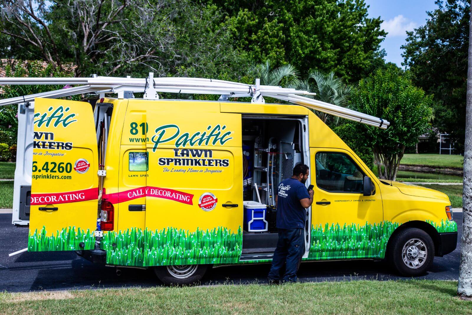 A yellow van with "Pacific Lawn Sprinklers" branding is parked with its side doors open. A person is standing near the open door, possibly working. Trees and a lawn are visible in the background.