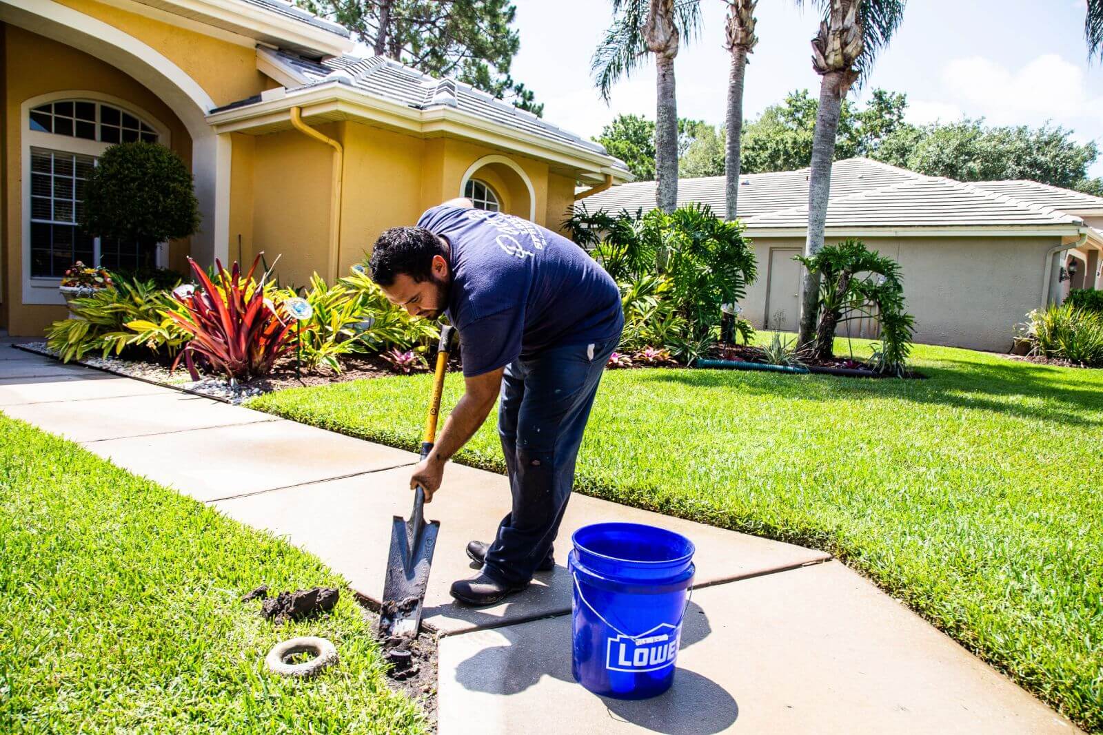 A man is working outdoors, digging a hole near a sidewalk in front of a house with a garden. A blue Lowe's bucket is placed nearby.