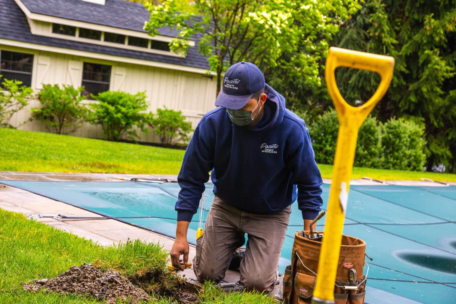 A worker in a blue hoodie and cap kneels on grass near a covered pool, using tools from a belt. A yellow shovel is in the foreground.