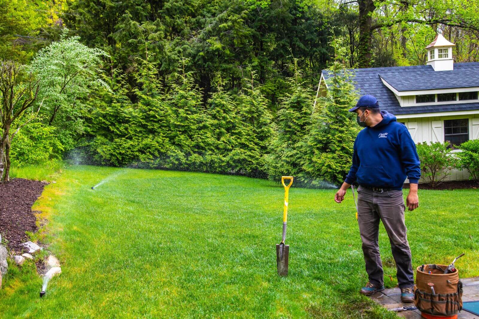 A man in a blue hoodie stands on a lawn next to a shovel stuck in the ground. He is observing a sprinkler watering the grass. A small white building and dense greenery are in the background.