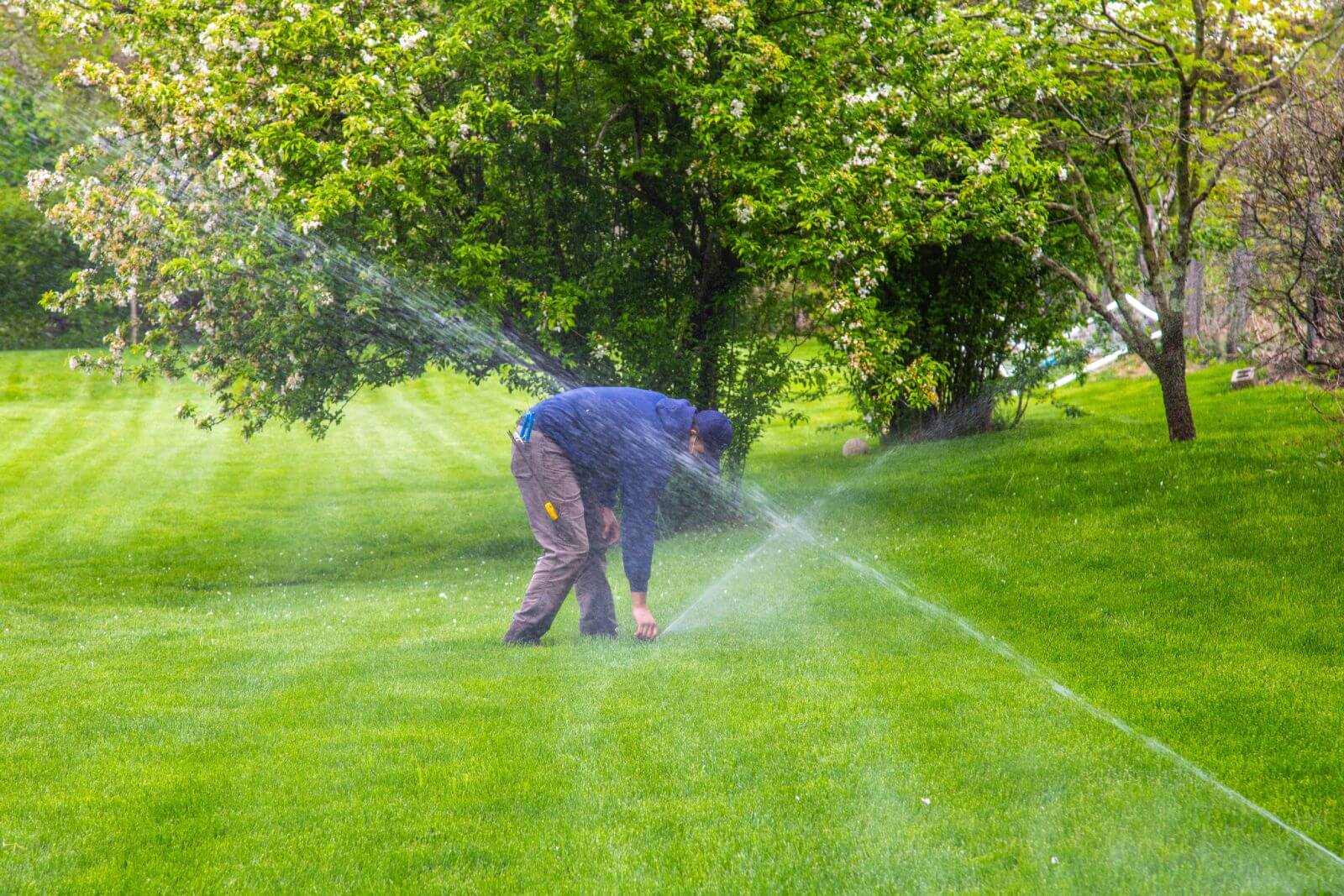 Person adjusting a sprinkler in a green, grassy area with trees in the background.