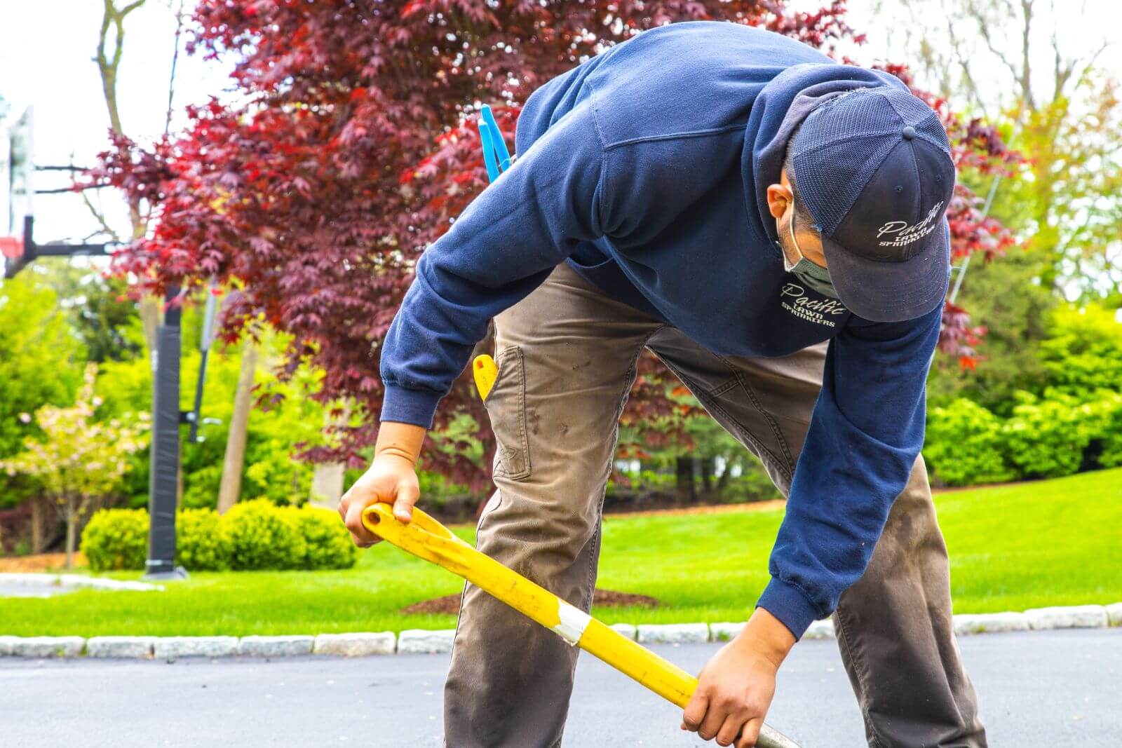 A worker in a blue sweatshirt and cap uses a yellow tool to perform outdoor maintenance on a paved surface, with greenery and a red-leaved tree in the background.