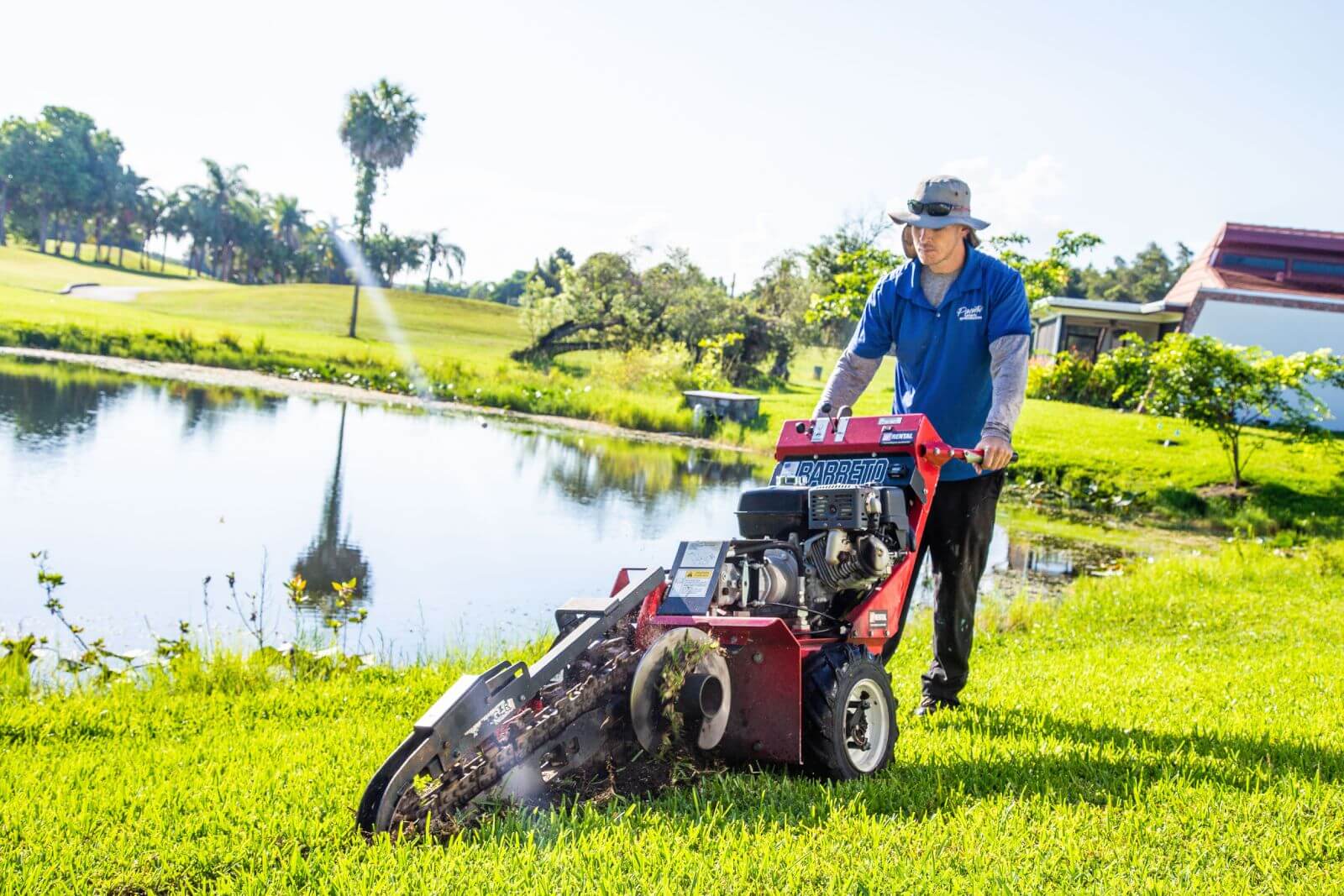 A person operates a red, wheeled machine on a grassy area beside a pond in a sunny outdoor setting.