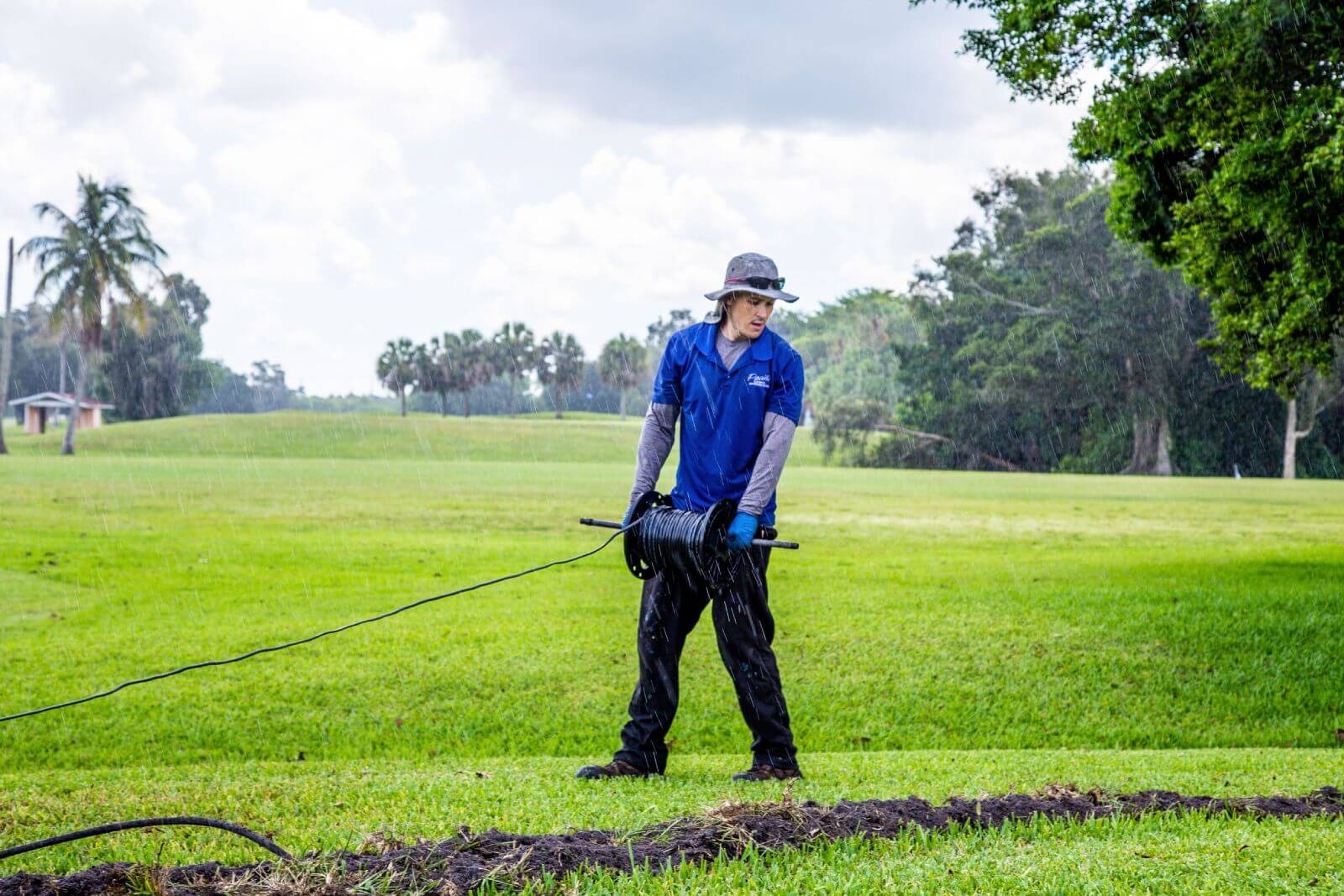 A person in a blue shirt and hat pulls a cable across a grassy area on a rainy day in an open field. Trees and cloudy sky are in the background.