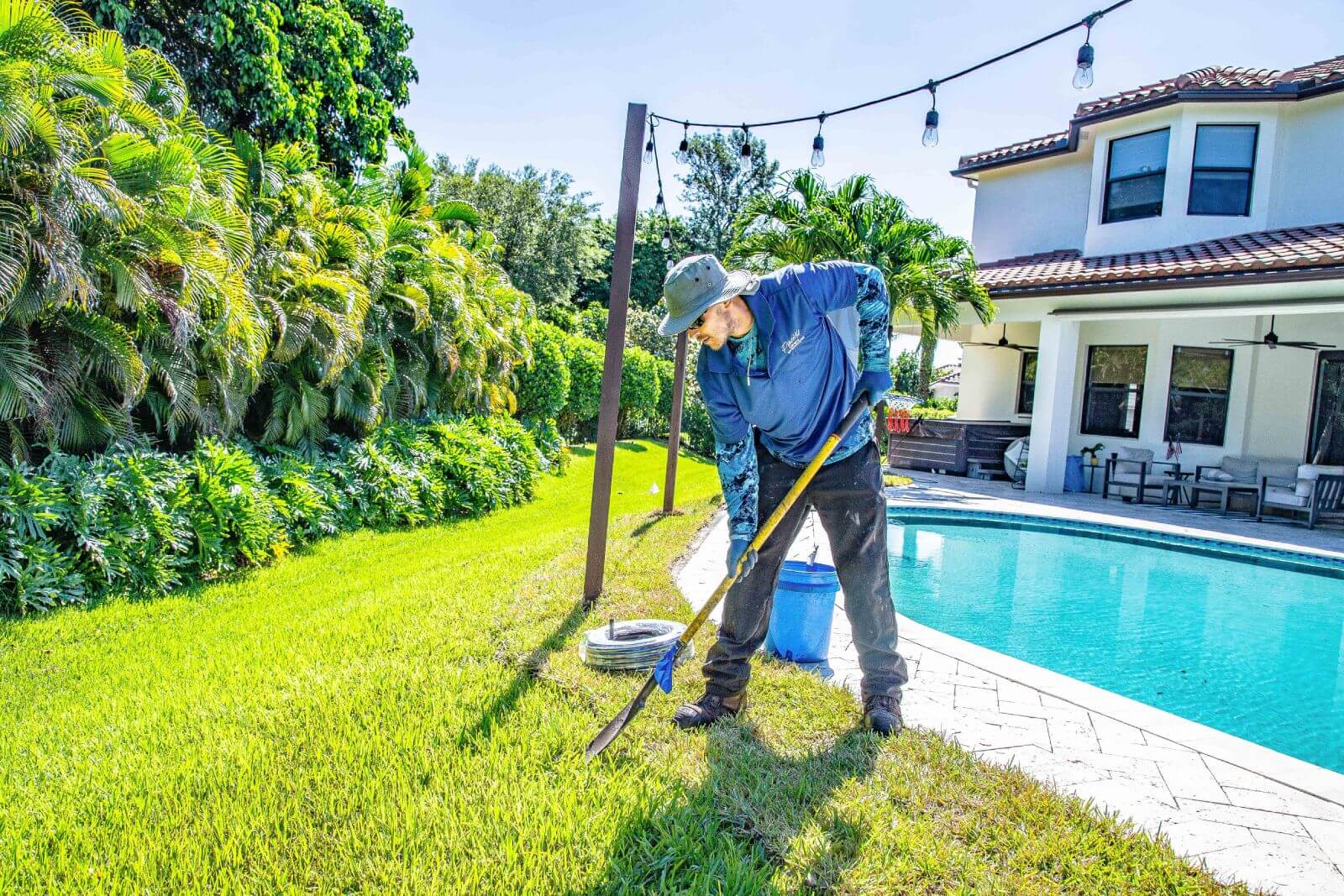 A person in a blue shirt and hat is digging in the grass near a swimming pool in a backyard with a white house and palm trees. They are using a shovel and appear to be working on a project.