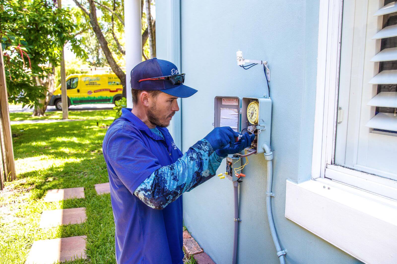 A technician in blue attire and gloves adjusts equipment inside an outdoor electrical box on the side of a light blue building. A yellow service van is parked in the background.