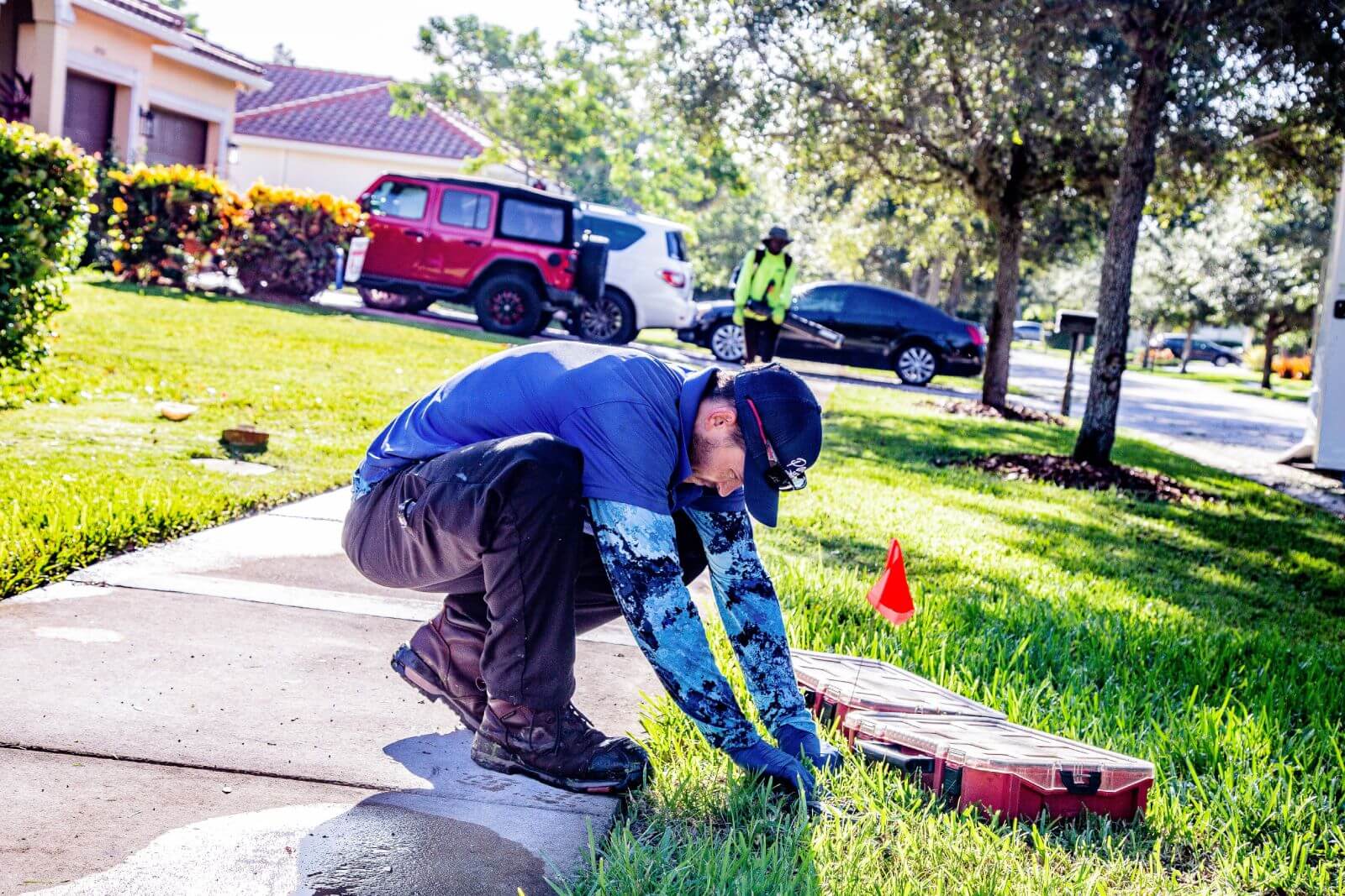 A man wearing a cap and long sleeves is crouching on a sidewalk, working with tools next to a red flag on a lawn. Another worker in a neon vest is in the background on the street.