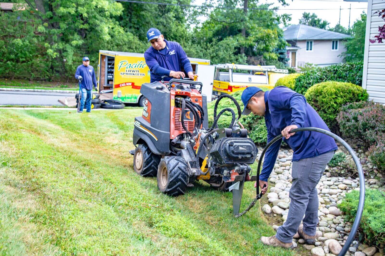 Workers operate a trenching machine and lay landscape pipes in a residential front yard with trees and trucks in the background.