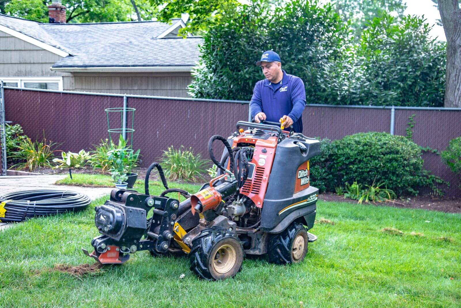 A person operates a trencher machine on a grassy lawn in a backyard, wearing protective gloves and a cap. A house, garden, and a fence are visible in the background.