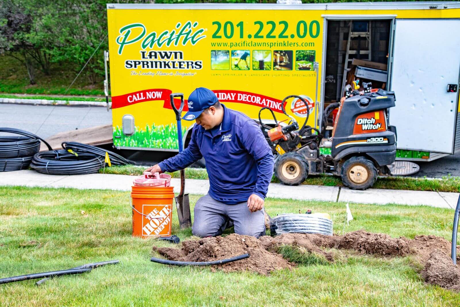 A worker in a blue uniform kneels on a lawn, handling tools and equipment for an irrigation system beside a Pacific Lawn Sprinklers trailer and a lawn mower.