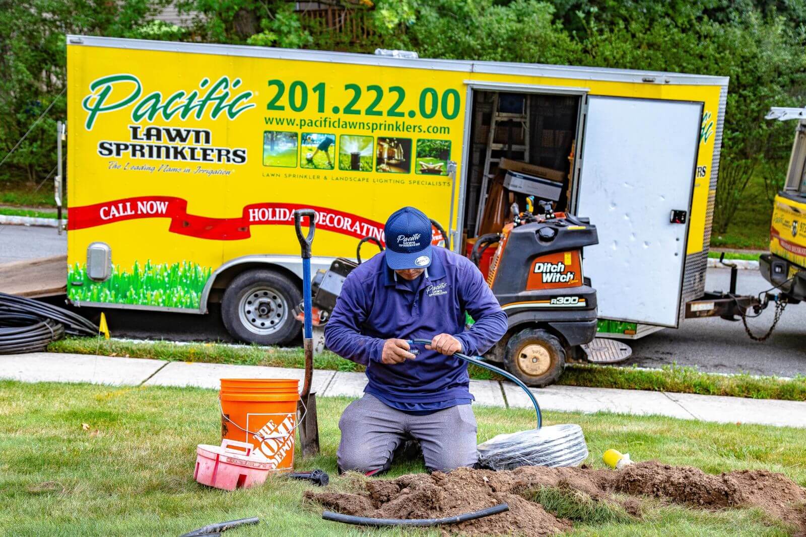 A worker kneels on a lawn to fix a sprinkler system in front of an open Pacific Lawn Sprinklers truck.