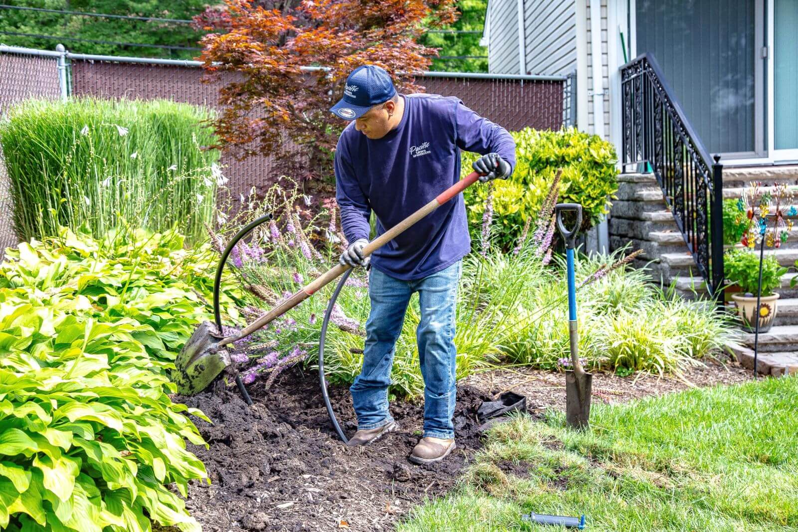 A person wearing a blue shirt and cap is using a shovel to work on a landscaped garden near a house. A spade is stuck in the ground beside them.