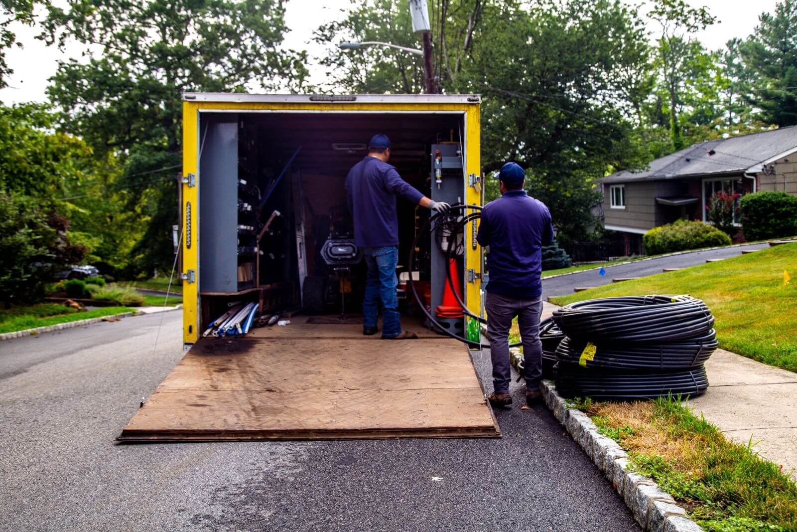 Two workers unloading equipment from the back of an open yellow truck parked on the street near a suburban neighborhood. One worker is handling cables. The area is surrounded by trees and houses.