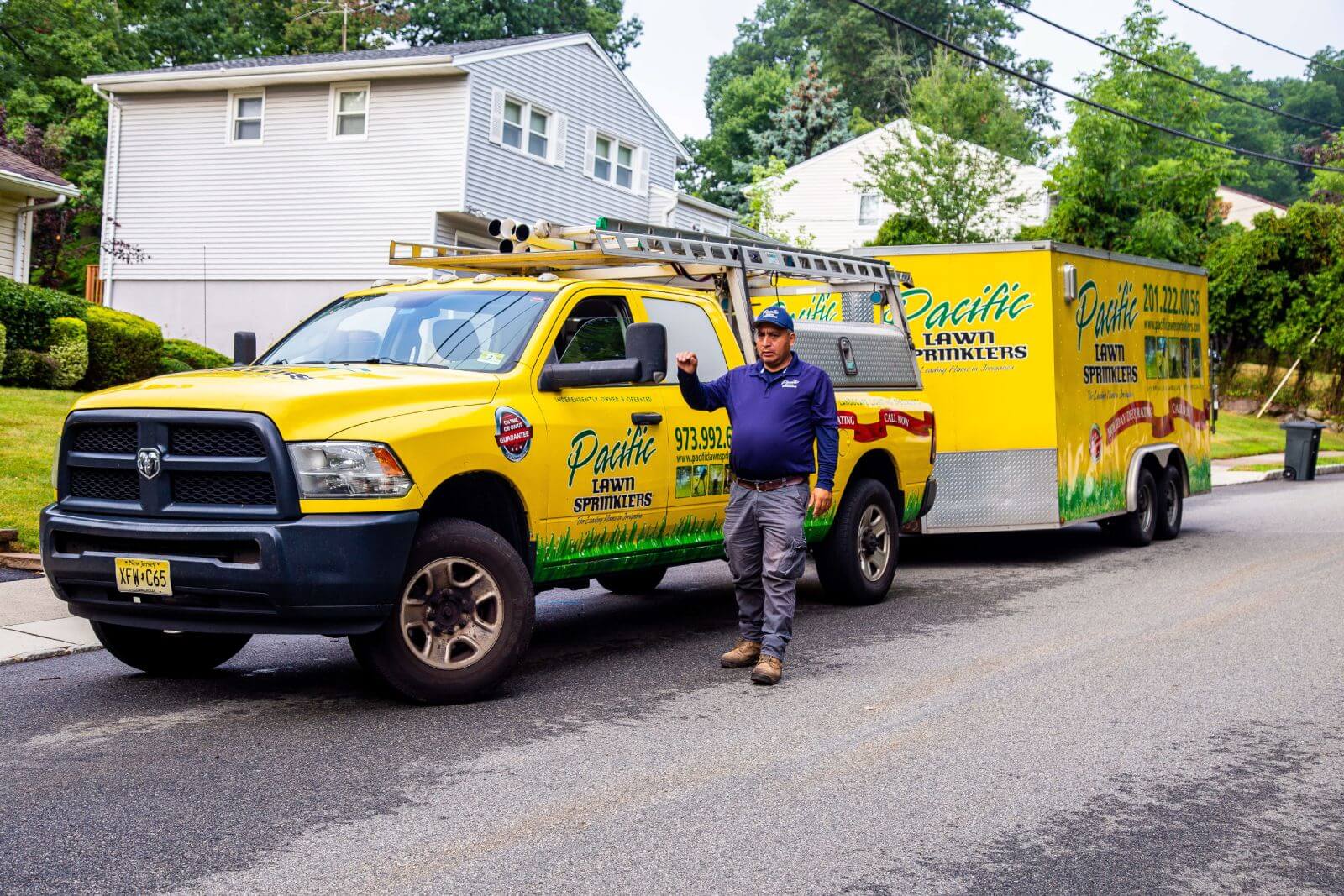 A worker stands beside a yellow Pacific Lawn Sprinklers truck with a trailer, parked on a residential street with houses and greenery in the background.