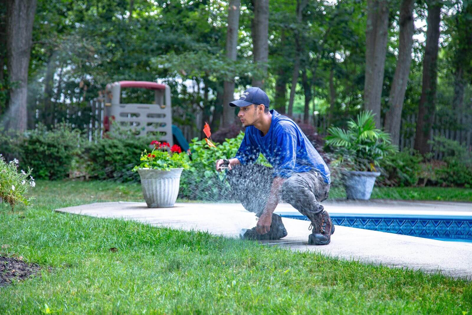 A person in a blue shirt is kneeling by the side of a swimming pool, using a hose to water the grass. There are potted plants and trees in the background.