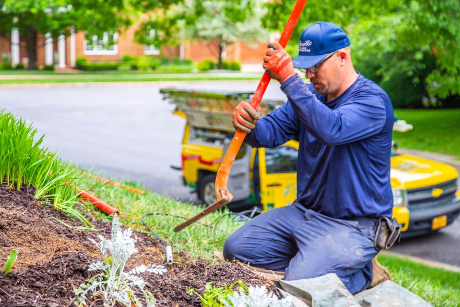 A worker in blue attire and an orange cap is using a manual post driver in a garden bed near a parked truck in a residential area.