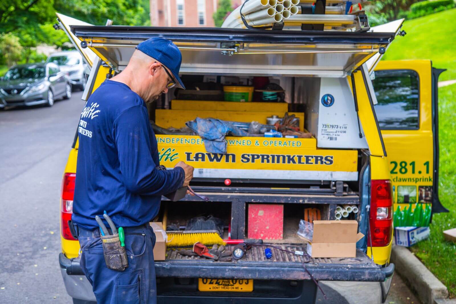 A worker in a blue uniform stands at the open rear of a yellow truck labeled "Lawn Sprinklers," sorting tools and materials. The truck's compartments are filled with various equipment and supplies.