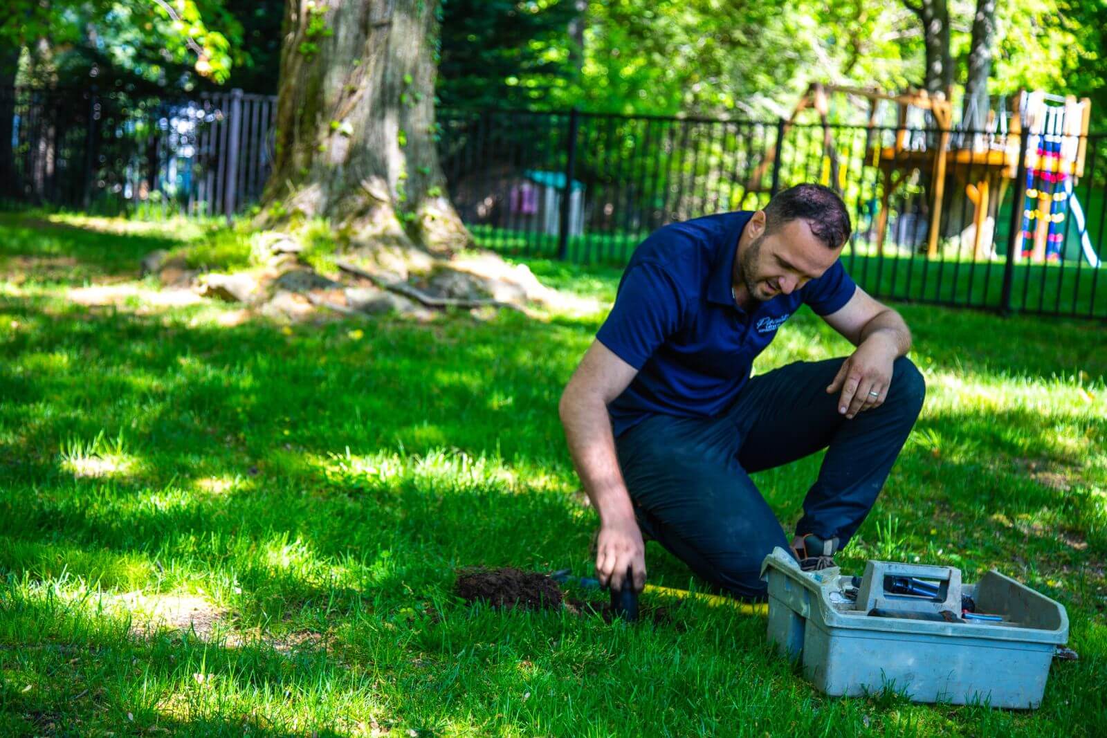 A man in a blue shirt is kneeling on the grass near a tree, working with a small shovel and tools from a box. A playground structure is visible in the background.