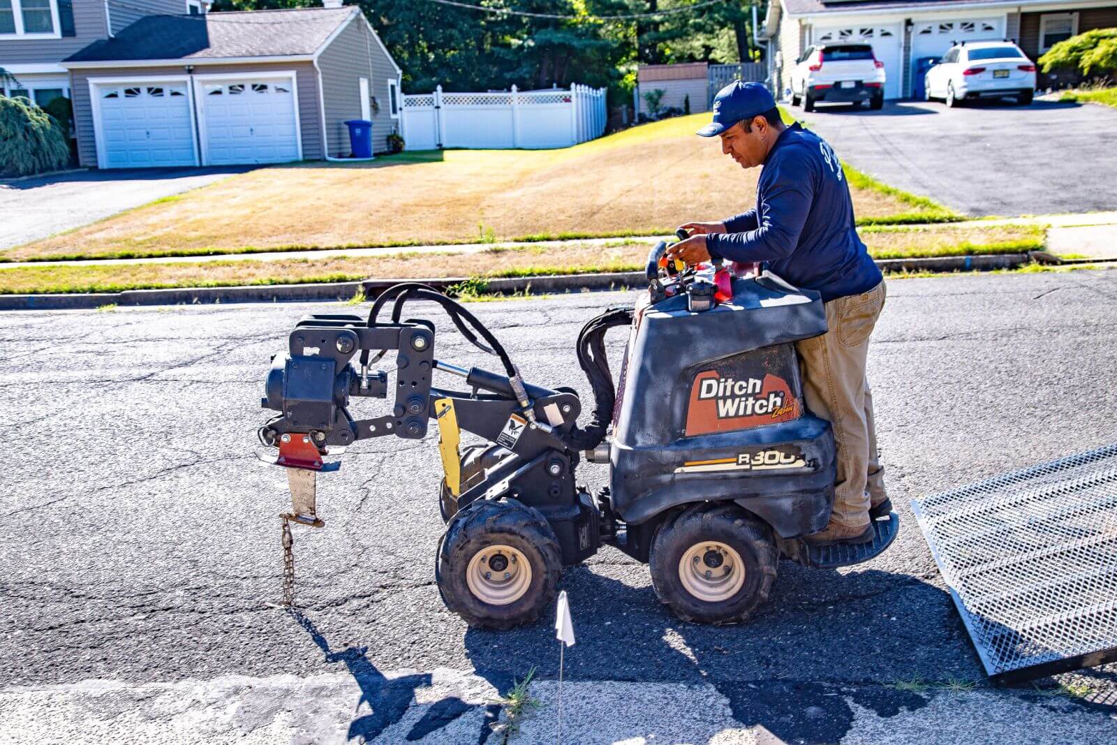 A worker operates a Ditch Witch R300 mini trencher on a residential street, cutting into asphalt. Two cars are parked in a driveway, and a ramp lies nearby.