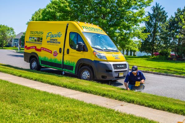 A person in a blue uniform is working on a lawn near a yellow Pacific Lawn Sprinklers van parked on a suburban street, showcasing the efficiency of franchise vs. own business approaches.