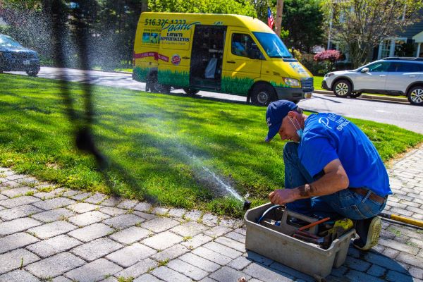 A man in a blue shirt and cap works on a small irrigation system on a paved pathway. A yellow service van is parked nearby on a residential street.