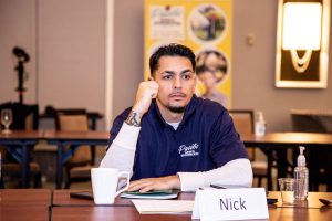 A man in a blue shirt sits at a conference table, confronting lawn care business challenges as he scribbles notes. In front of him are a notepad, cup, hand sanitizer, and a nameplate bearing "Nick.