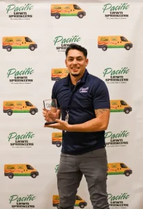 Person holding a plaque in front of a backdrop with "Pacific Lawn Sprinklers" logos and images of trucks.