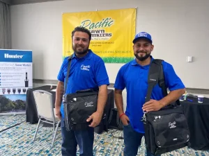 Two men in blue shirts and hats, smiling and holding black bags, stand in front of a banner for Pacific Lawn Sprinklers at an indoor event.