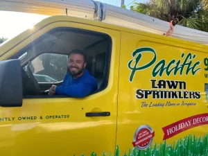 A man sitting in the driver's seat of a bright yellow van with "Pacific Lawn Sprinklers" text on the side.