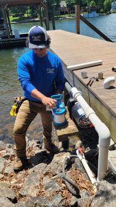 A person in blue attire, likely facing lawn care business challenges, works on a water pump system by a lakeside dock. Wearing a cap, they adjust the equipment surrounded by pipes, rocks, and a wooden structure.