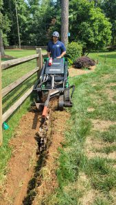 A person operates a trencher machine, expertly digging a narrow trench along a grassy area next to a wooden fence. Surrounded by lush greenery and woods, this task highlights some of the lawn care business challenges faced in maintaining pristine landscapes.