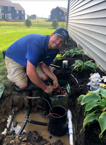 A man in a blue shirt kneels by a house, addressing wiring and pipes in an outdoor setting with plants nearby. His focus mirrors the meticulous attention needed to navigate lawn care business challenges successfully.