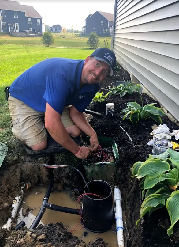 A person in a blue shirt and cap is working on outdoor wiring near a house, surrounded by plants and soil.